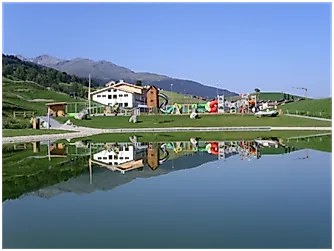 Spiegelung eines Bergspielplatzes mit Gebäuden im klaren Bergsee am Fuße von Surcuolm – Piz Mundaun an einem sonnigen Sommertag.