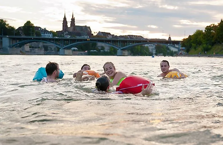 Junge Leute treiben beim beliebten Rheinschwimmen mit bunten Schweimmtieren durch Basel mit Altstadt im Hintergrund
